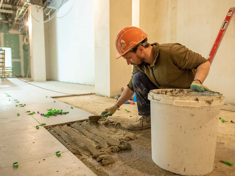 Profissional de capacete aplicando argamassa no piso para o assentamento de revestimento — imagem ilustrativa.